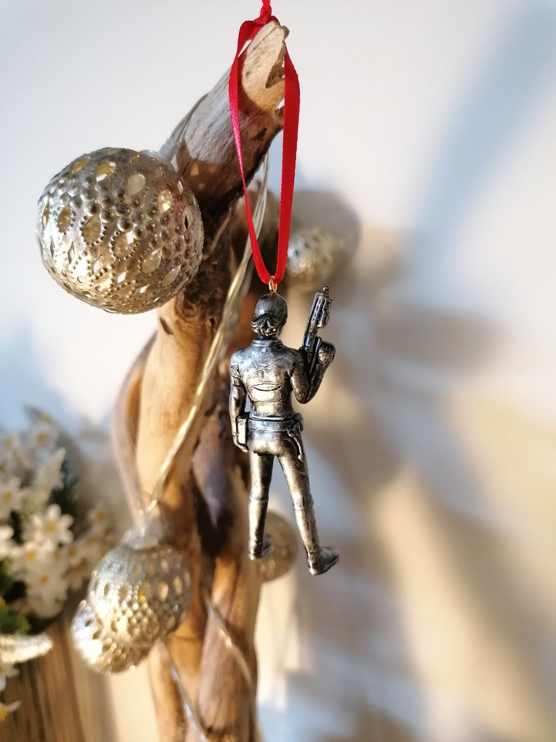 May include: A silver-coloured figurine of a person holding a weapon, hanging from a red ribbon on a branch. The figurine is a Christmas ornament.
