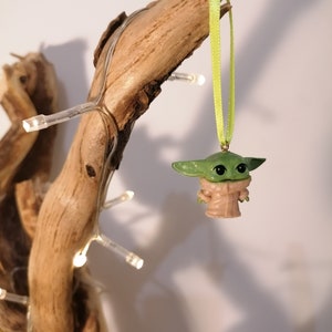 May include: A green and tan Baby Yoda ornament hanging from a light green ribbon. The ornament is suspended from a piece of driftwood, with small white lights wrapped around it. The background is a plain white.