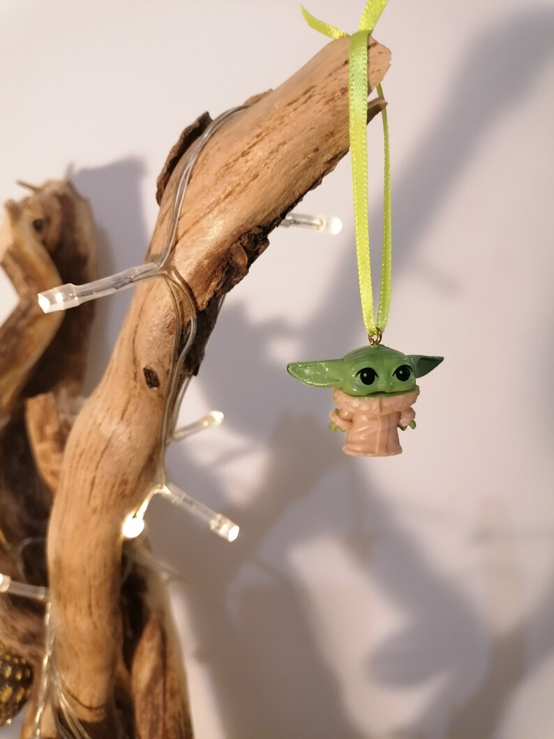 May include: A green and tan Baby Yoda ornament hanging from a light green ribbon. The ornament is suspended from a piece of driftwood, with small white lights wrapped around it. The background is a plain white.