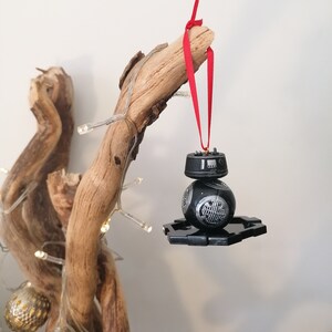 K&ouml;nnte beinhalten: Ein schwarzer und silberner Star Wars BB-8 Droiden-Weihnachtsbaumschmuck, der an einem roten Band an einem Treibholzast h&auml;ngt.