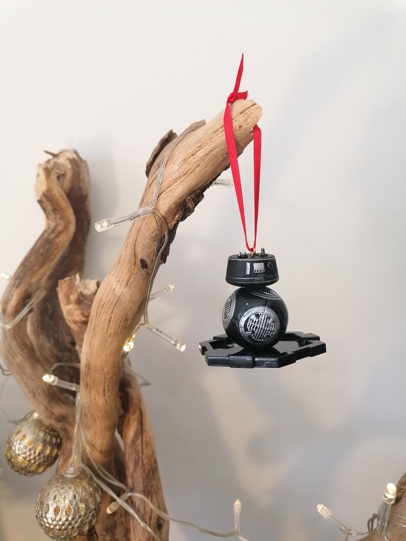 K&ouml;nnte beinhalten: Ein schwarzer und silberner Star Wars BB-8 Droiden-Weihnachtsbaumschmuck, der an einem roten Band an einem Treibholzast h&auml;ngt.