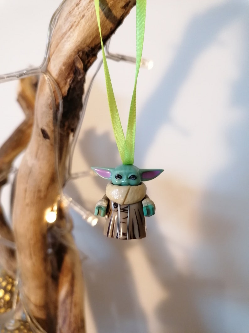 Puede incluir: Un adorno de personaje verde y marr&oacute;n con orejas grandes, que lleva un abrigo marr&oacute;n con cuello de piel. El adorno est&aacute; colgado de una cinta verde.
