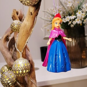 May include: A plastic doll ornament of a princess wearing a blue dress with a pink cape and a crown. The doll is hanging from a purple string.