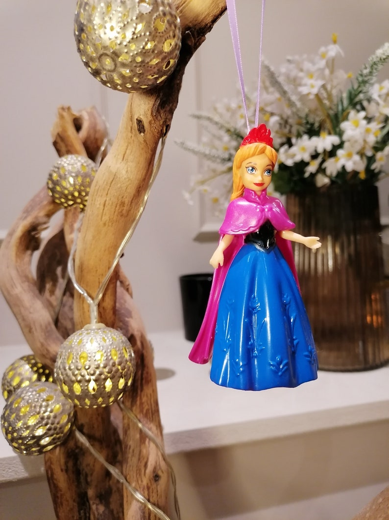 May include: A plastic doll ornament of a princess wearing a blue dress with a pink cape and a crown. The doll is hanging from a purple string.