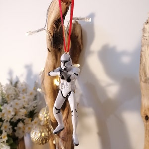 Puede incluir: Un adorno navide&ntilde;o de pl&aacute;stico blanco con forma de Stormtrooper de Star Wars colgado de una cinta roja en un trozo de madera a la deriva.