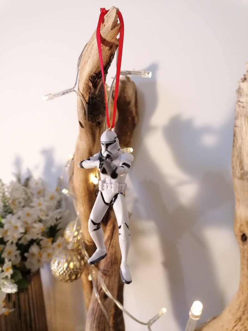 Puede incluir: Un adorno navide&ntilde;o de pl&aacute;stico blanco con forma de Stormtrooper de Star Wars colgado de una cinta roja en un trozo de madera a la deriva.
