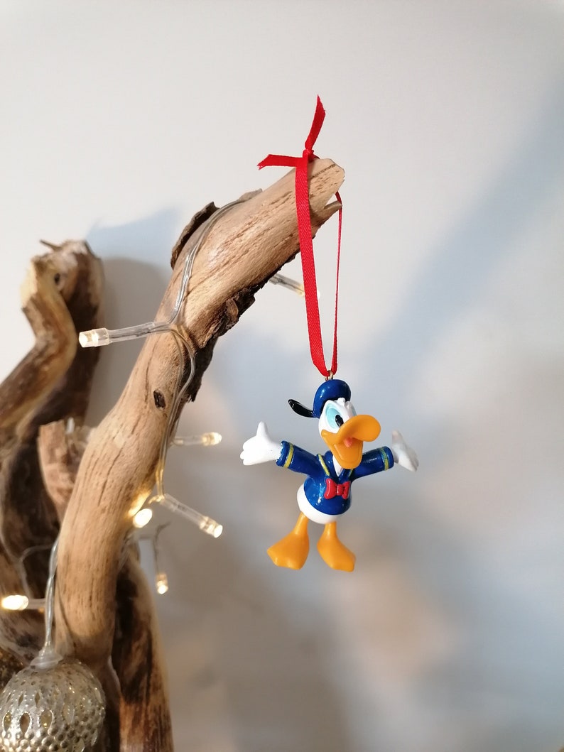 May include: A yellow and blue plastic Donald Duck Christmas ornament with a red bow hanging from a driftwood branch with white lights.