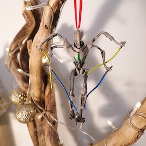 May include: A gray and green robot Christmas ornament with red string hanging from a branch. The robot has four arms and is holding two blue and yellow lights.