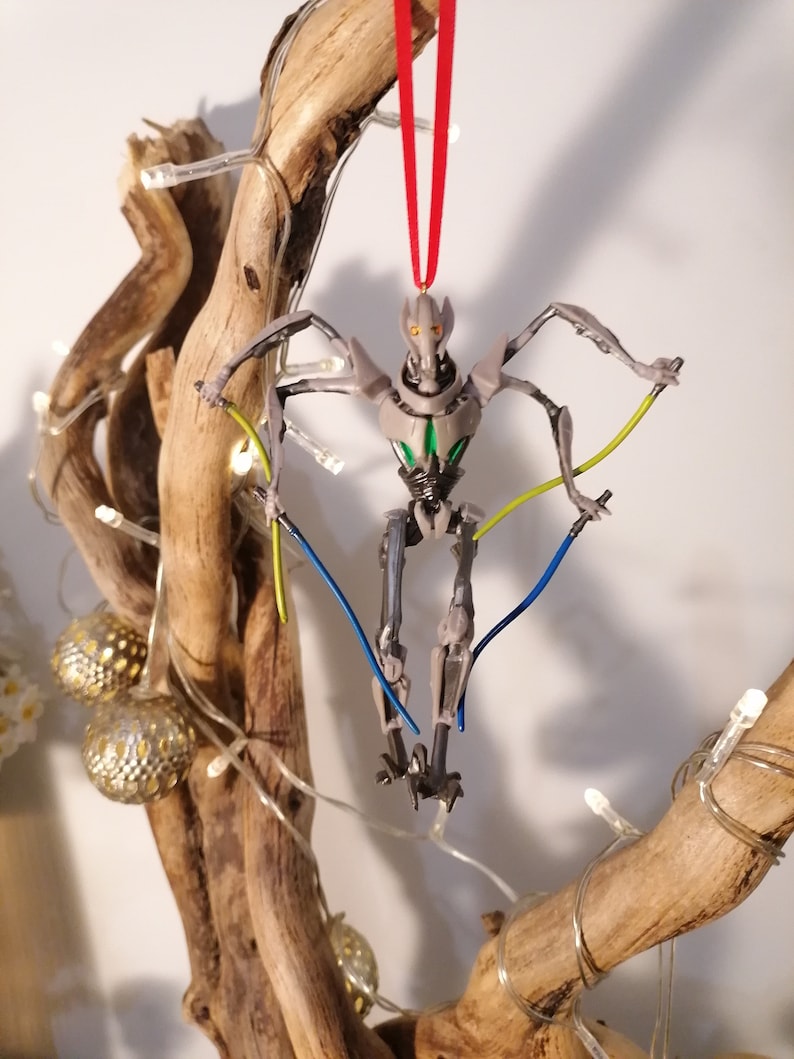 May include: A gray and green robot Christmas ornament with red string hanging from a branch. The robot has four arms and is holding two blue and yellow lights.