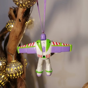 Peut inclure: Ornement de No&euml;l Buzz l'&Eacute;clair, aux couleurs blanc, vert et violet. La figurine jouet a des ailes d&eacute;ploy&eacute;es et est suspendue par un ruban violet. L'ornement est suspendu devant une branche en bois avec des sph&egrave;res d&eacute;coratives illumin&eacute;es.