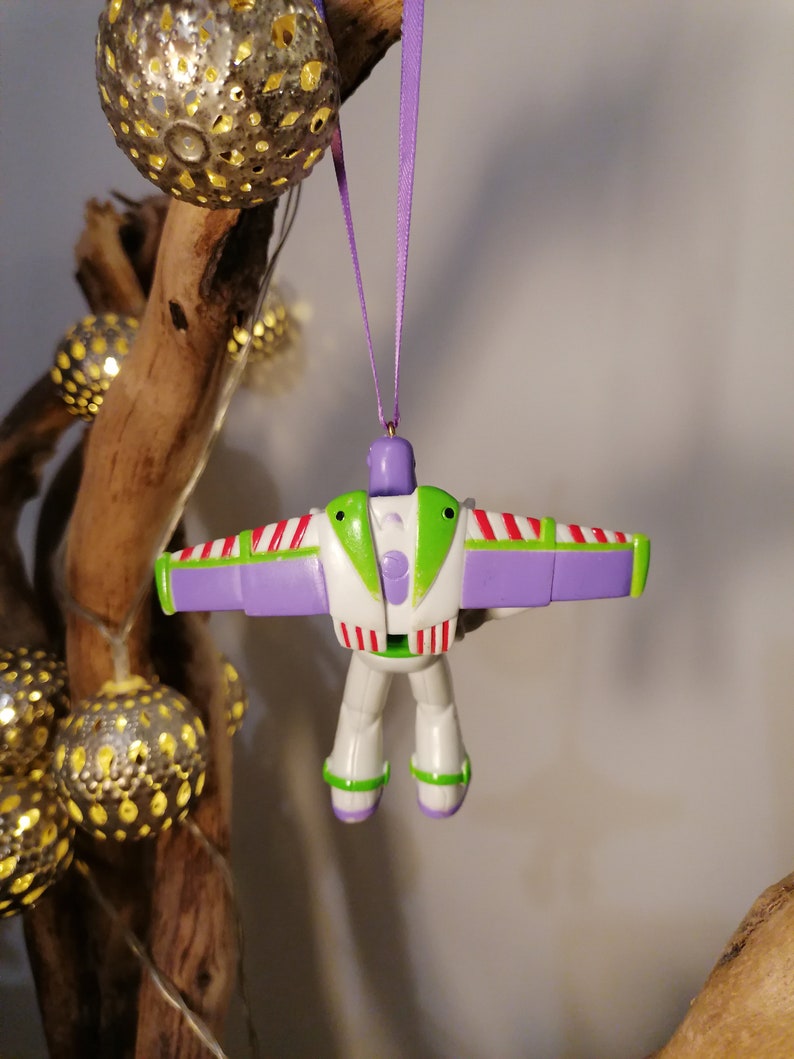 Peut inclure: Ornement de No&euml;l Buzz l'&Eacute;clair, aux couleurs blanc, vert et violet. La figurine jouet a des ailes d&eacute;ploy&eacute;es et est suspendue par un ruban violet. L'ornement est suspendu devant une branche en bois avec des sph&egrave;res d&eacute;coratives illumin&eacute;es.