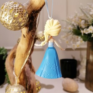 May include: A blue and white plastic ornament featuring a small doll with blonde hair and a blue dress. The doll is wearing a blue dress with white snowflake designs. The ornament is hanging from a blue ribbon.
