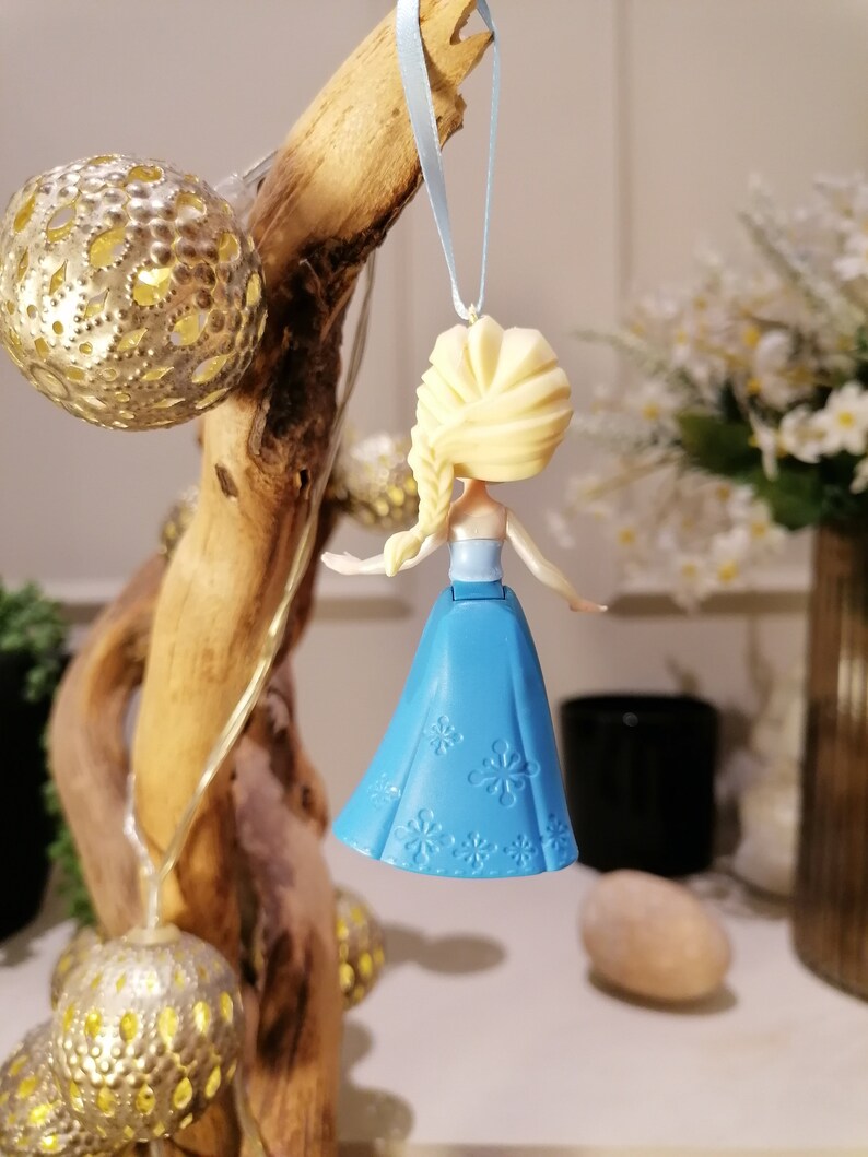 May include: A blue and white plastic ornament featuring a small doll with blonde hair and a blue dress. The doll is wearing a blue dress with white snowflake designs. The ornament is hanging from a blue ribbon.