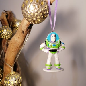 Puede incluir: Una cinta morada cuelga de una rama con una figura de pl&aacute;stico blanca y verde de Buzz Lightyear de la serie de pel&iacute;culas Toy Story. La figura lleva un uniforme de ranger espacial y tiene un casco azul con visera. La figura cuelga de una rama con una cadena de luces.
