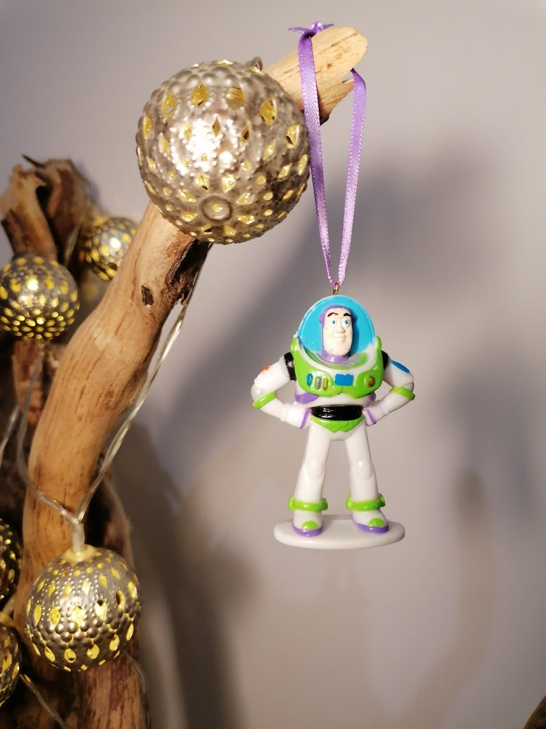Puede incluir: Una cinta morada cuelga de una rama con una figura de pl&aacute;stico blanca y verde de Buzz Lightyear de la serie de pel&iacute;culas Toy Story. La figura lleva un uniforme de ranger espacial y tiene un casco azul con visera. La figura cuelga de una rama con una cadena de luces.