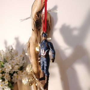 May include: A blue and red Captain America figurine ornament hanging from a red ribbon on a piece of driftwood.