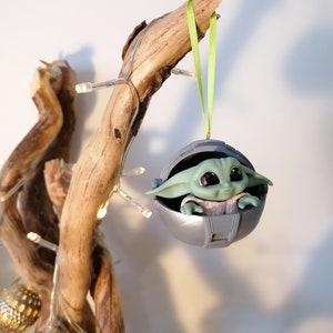 May include: A green and gray Christmas ornament featuring a character from the Star Wars franchise, The Mandalorian. The character is sitting in a pod-like structure with a green ribbon attached.