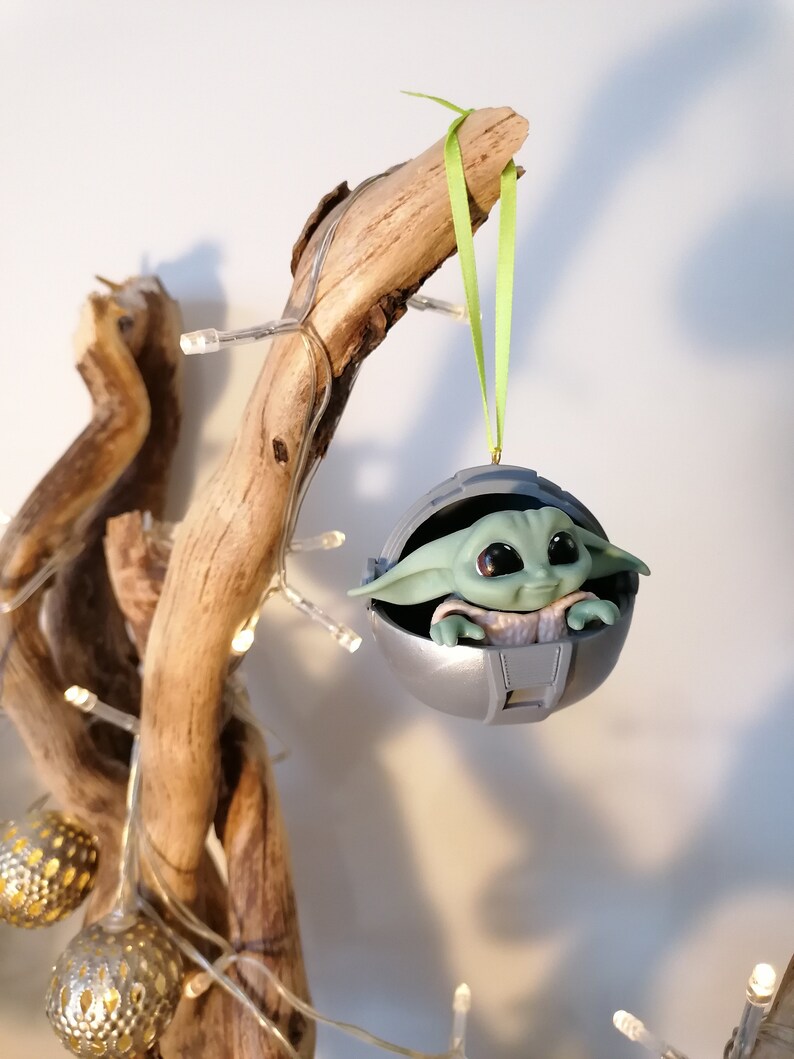 May include: A green and gray Christmas ornament featuring a character from the Star Wars franchise, The Mandalorian. The character is sitting in a pod-like structure with a green ribbon attached.