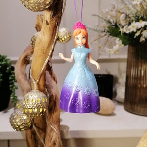K&ouml;nnte beinhalten: Ein lila und blauer Prinzessinnen-Puppen-Anh&auml;nger mit einer rosa Krone, der an einer lila Schnur h&auml;ngt. Die Puppe tr&auml;gt ein hellblaues Kleid mit einem lila Ombre-Effekt und Schneeflocken-Details.