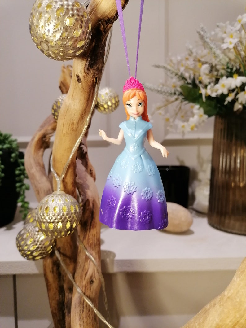 K&ouml;nnte beinhalten: Ein lila und blauer Prinzessinnen-Puppen-Anh&auml;nger mit einer rosa Krone, der an einer lila Schnur h&auml;ngt. Die Puppe tr&auml;gt ein hellblaues Kleid mit einem lila Ombre-Effekt und Schneeflocken-Details.