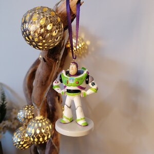 May include: A green and white plastic figurine of Buzz Lightyear from the Toy Story franchise hanging from a purple ribbon. The figurine is wearing a space ranger uniform with a green vest and a white helmet. The figurine is hanging from a branch with a string of lights.