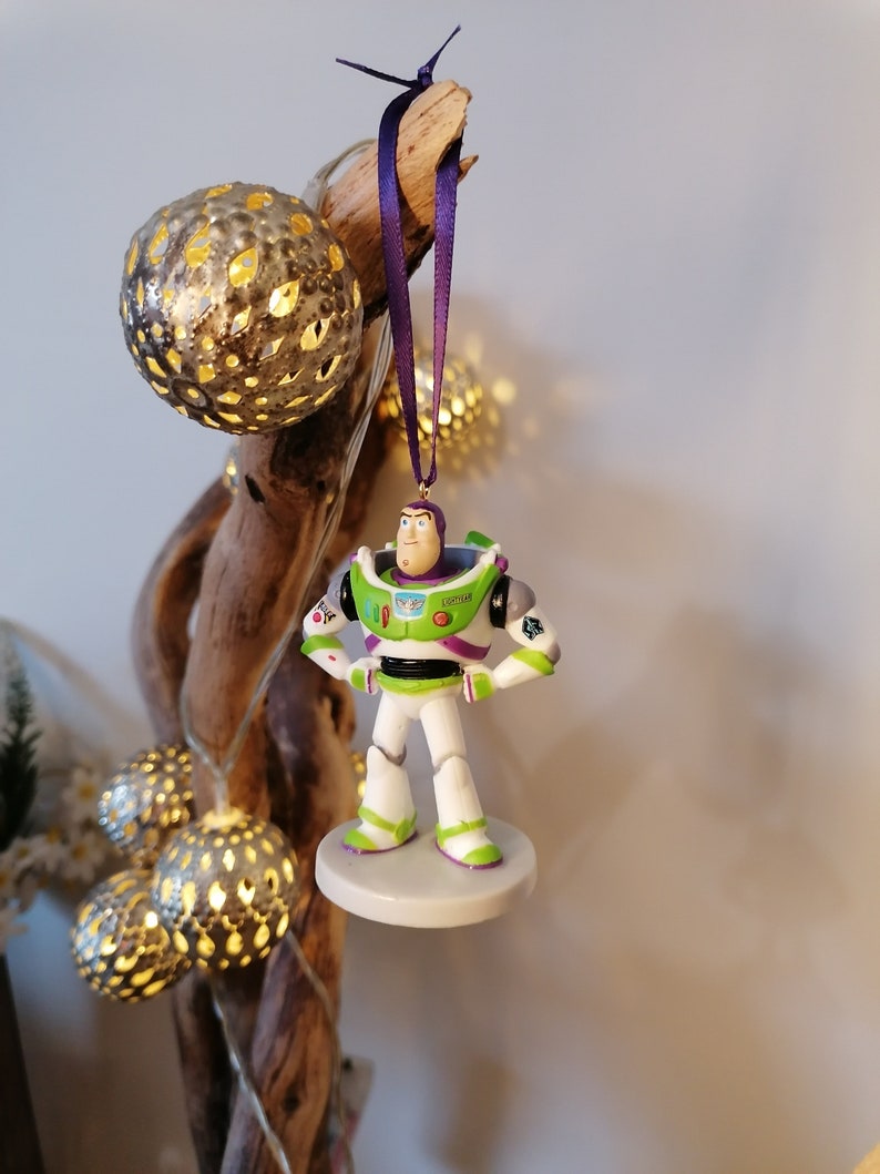 May include: A green and white plastic figurine of Buzz Lightyear from the Toy Story franchise hanging from a purple ribbon. The figurine is wearing a space ranger uniform with a green vest and a white helmet. The figurine is hanging from a branch with a string of lights.