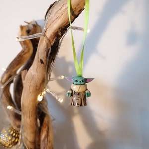Puede incluir: Una figurita verde y marr&oacute;n de un personaje de la franquicia Star Wars, colgada de una cinta verde en una rama de madera flotante. La figurita es una figura peque&ntilde;a, infantil, con orejas grandes y una t&uacute;nica marr&oacute;n.