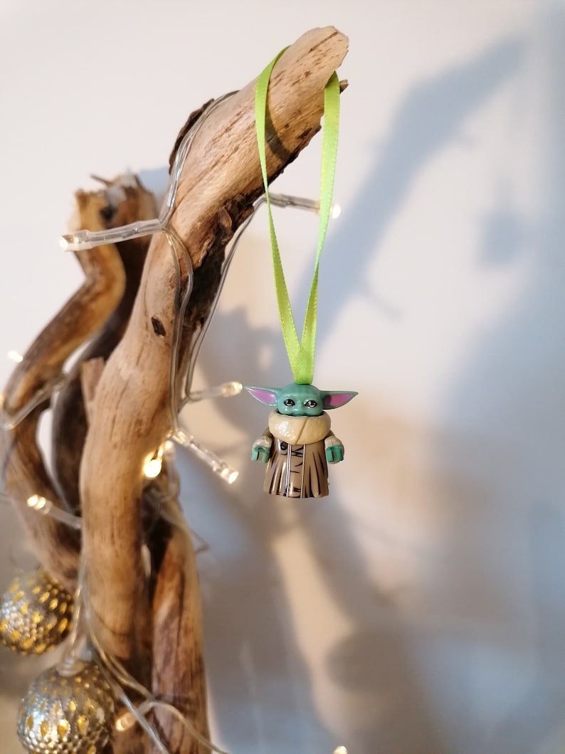 Puede incluir: Una figurita verde y marr&oacute;n de un personaje de la franquicia Star Wars, colgada de una cinta verde en una rama de madera flotante. La figurita es una figura peque&ntilde;a, infantil, con orejas grandes y una t&uacute;nica marr&oacute;n.