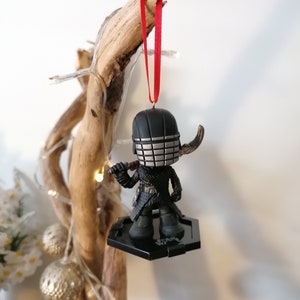 May include: Black and silver Star Wars ornament featuring a character with a helmet and a lightsaber. The ornament is hanging from a red ribbon on a wooden branch.