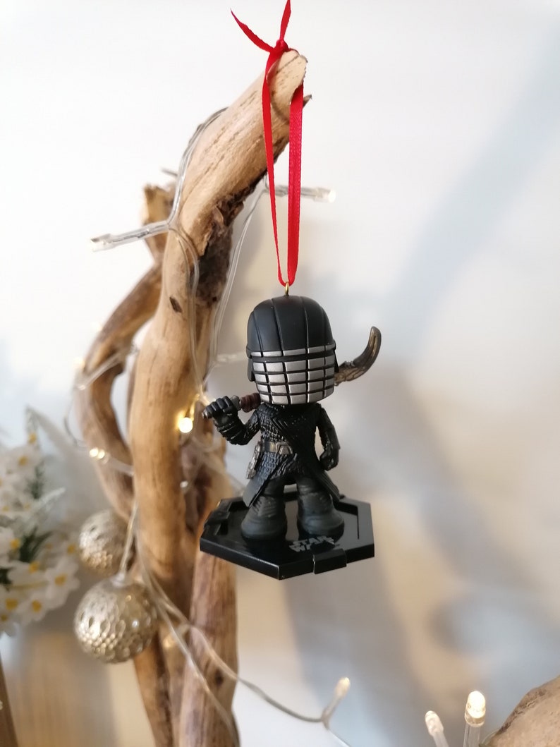 May include: Black and silver Star Wars ornament featuring a character with a helmet and a lightsaber. The ornament is hanging from a red ribbon on a wooden branch.