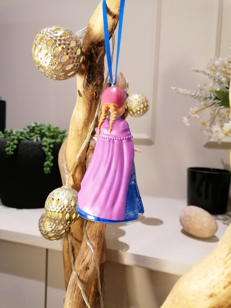 Puede incluir: Un adorno de pl&aacute;stico morado y azul de una ni&ntilde;a con el pelo largo y casta&ntilde;o, que lleva una capa morada y un vestido azul. El adorno est&aacute; colgado de una cinta azul en una rama de madera.