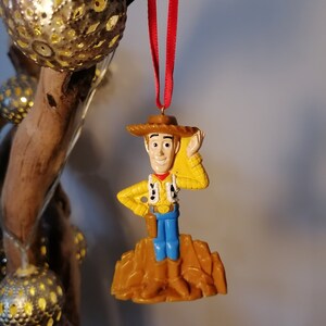 May include: A plastic ornament of Woody from Toy Story. He is wearing his signature cowboy hat and vest, and is standing on a brown base. He is holding his right hand up to his hat, as if he is tipping it.