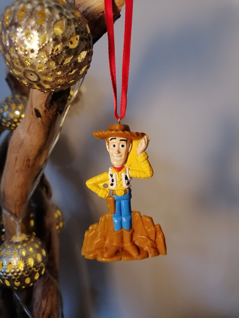 May include: A plastic ornament of Woody from Toy Story. He is wearing his signature cowboy hat and vest, and is standing on a brown base. He is holding his right hand up to his hat, as if he is tipping it.