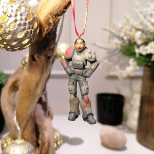 Puede incluir: Una figurita de plástico gris y rojo de una persona con casco y armadura, colgada de una cuerda roja. La figurita es un adorno navideño.