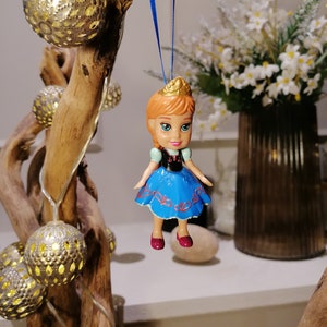 K&ouml;nnte beinhalten: Eine kleine Plastikpuppe mit einem blauen Kleid und roten Schuhen h&auml;ngt an einer blauen Schnur. Die Puppe tr&auml;gt eine Krone auf dem Kopf und lange orange Haare. Die Puppe h&auml;ngt an einem Ast mit wei&szlig;en Lichtern, die darum gewickelt sind.