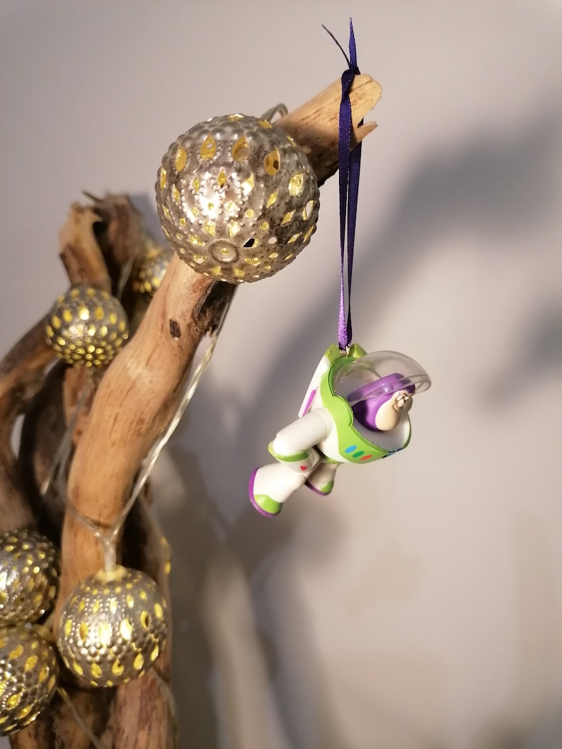May include: A green and white astronaut toy ornament hanging from a branch with a purple ribbon. The ornament is surrounded by gold, metallic, sphere-shaped ornaments.