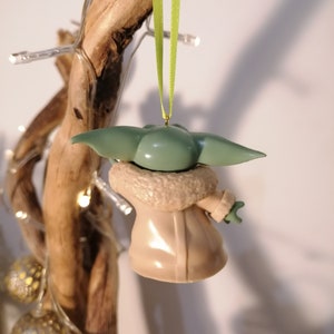 May include: A green and brown Christmas ornament shaped like a character from the Star Wars franchise. The ornament is hanging from a green ribbon.