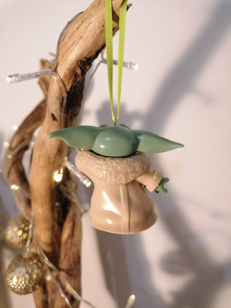 May include: A green and brown Christmas ornament shaped like a character from the Star Wars franchise. The ornament is hanging from a green ribbon.