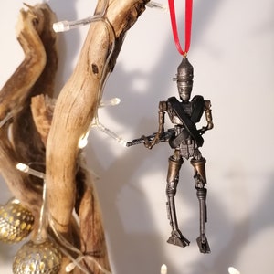 Puede incluir: Un adorno de robot de color plateado y bronce con una cinta roja colgando de una rama de madera. El robot sostiene un arma y tiene una correa negra cruzada sobre su pecho.