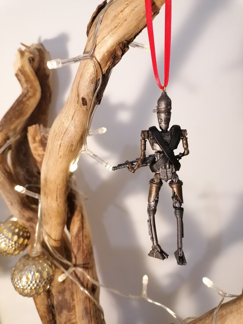 Puede incluir: Un adorno de robot de color plateado y bronce con una cinta roja colgando de una rama de madera. El robot sostiene un arma y tiene una correa negra cruzada sobre su pecho.