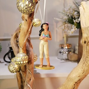 May include: A figurine of a woman with long dark hair wearing a blue and white top and a beige skirt. She is holding a pink flower and is standing on a yellow base. The figurine is hanging from a white string on a branch with string lights.
