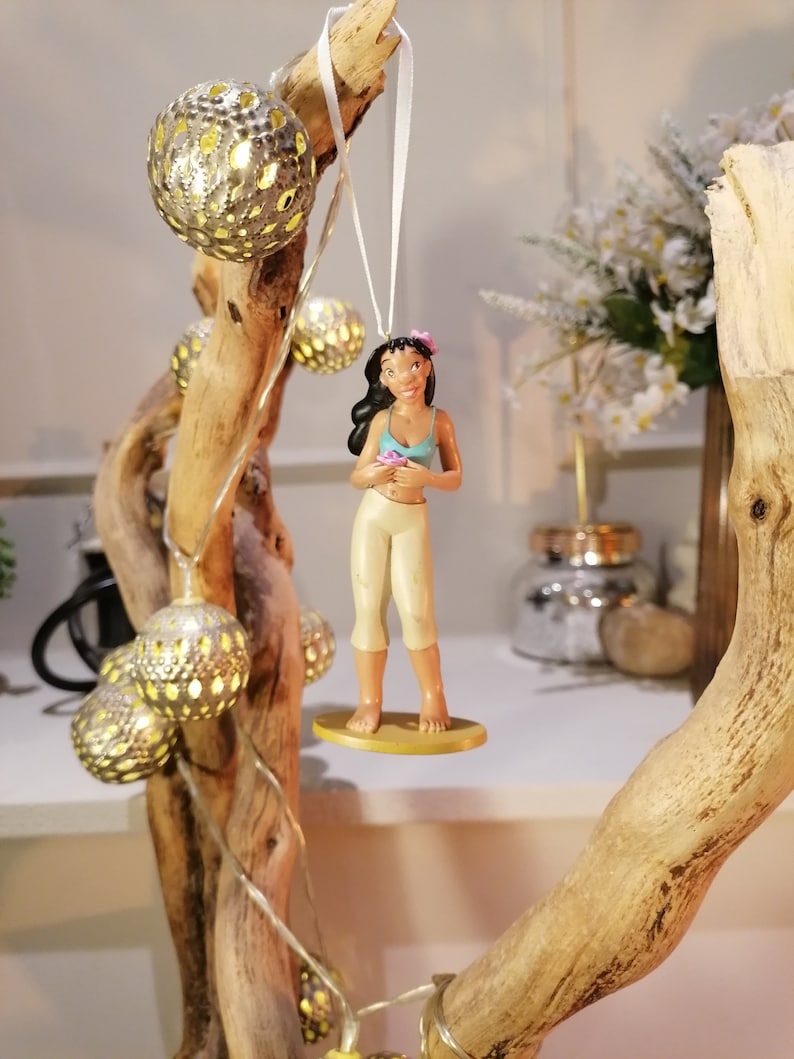May include: A figurine of a woman with long dark hair wearing a blue and white top and a beige skirt. She is holding a pink flower and is standing on a yellow base. The figurine is hanging from a white string on a branch with string lights.