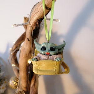 May include: A green and yellow Christmas ornament featuring a character from the Star Wars franchise, The Mandalorian. The character is sitting in a yellow bag with a green ribbon.