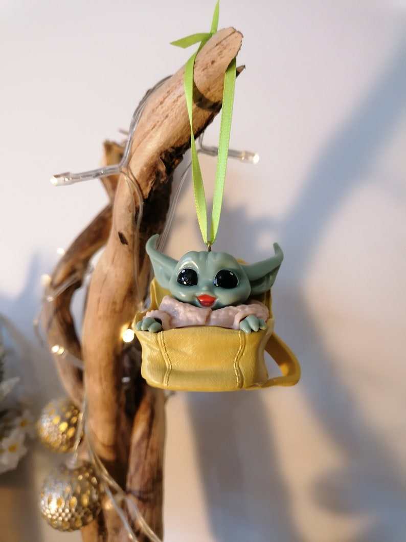 May include: A green and yellow Christmas ornament featuring a character from the Star Wars franchise, The Mandalorian. The character is sitting in a yellow bag with a green ribbon.