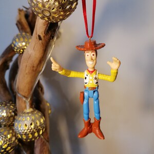 May include: A plastic toy ornament of Woody from Toy Story. He is wearing his signature cowboy hat, vest, and jeans. He is holding his arm out, as if he is pointing. The ornament is hanging from a red ribbon.