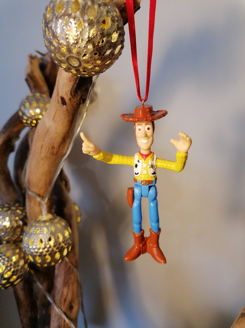 May include: A plastic toy ornament of Woody from Toy Story. He is wearing his signature cowboy hat, vest, and jeans. He is holding his arm out, as if he is pointing. The ornament is hanging from a red ribbon.
