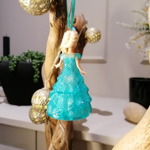May include: A blue plastic ornament shaped like a princess in a sparkly blue dress. The ornament is hanging from a branch with a blue ribbon.