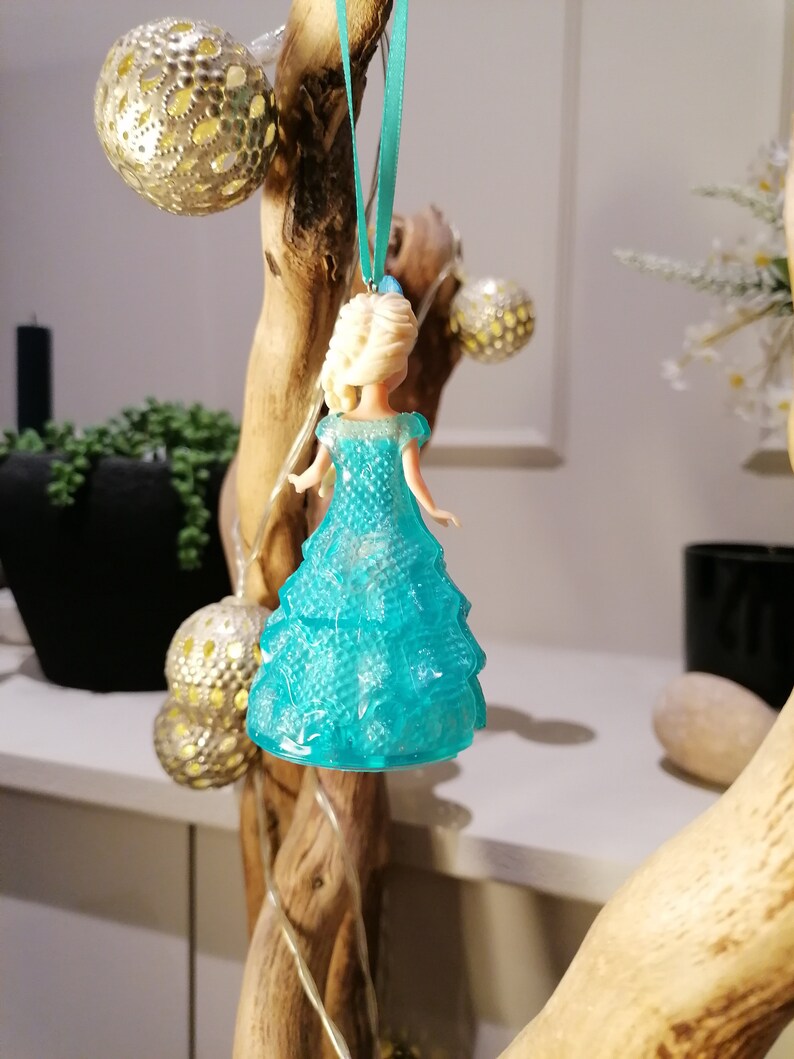 May include: A blue plastic ornament shaped like a princess in a sparkly blue dress. The ornament is hanging from a branch with a blue ribbon.
