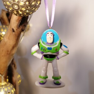 Puede incluir: Un adorno de pl&aacute;stico verde y blanco de Buzz Lightyear de la franquicia Toy Story. El adorno est&aacute; colgado de una cinta morada.