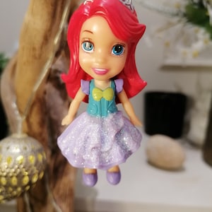 Peut inclure: Une petite poup&eacute;e en plastique d'Ariel de La Petite Sir&egrave;ne portant une robe violette &agrave; paillettes, un corsage turquoise et une couronne argent&eacute;e. Elle a les cheveux roux et les yeux bleus.